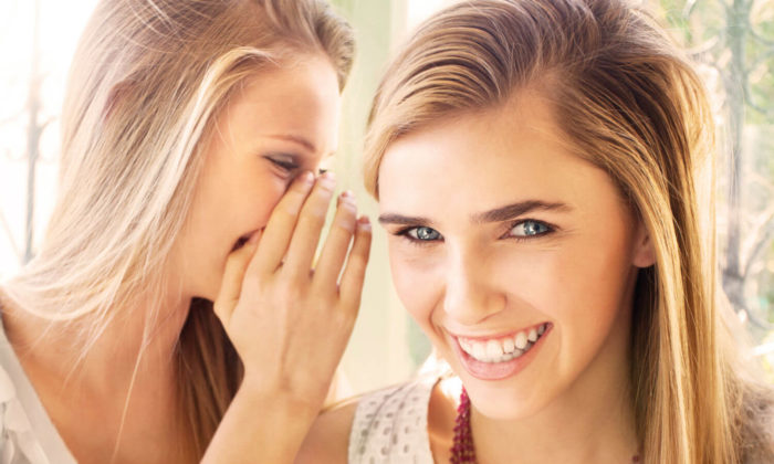 Headshot of two blond women. One is wishering into the other ear while she is smiling at the camera.