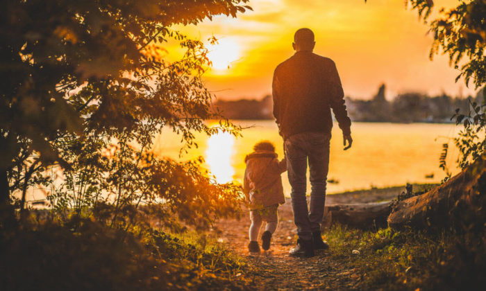 Man and small child walking into the sunset with a body of water in front of them