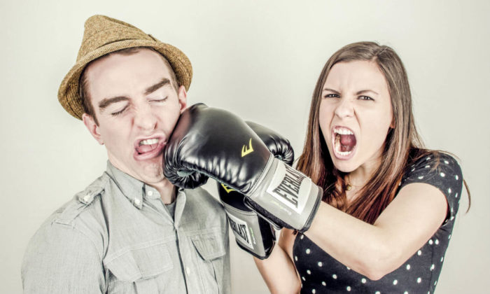 A girl has boxing gloves on is punching a man in the face. She looks ferocious. The man has a hat on and is making a funny face in response to being punched.