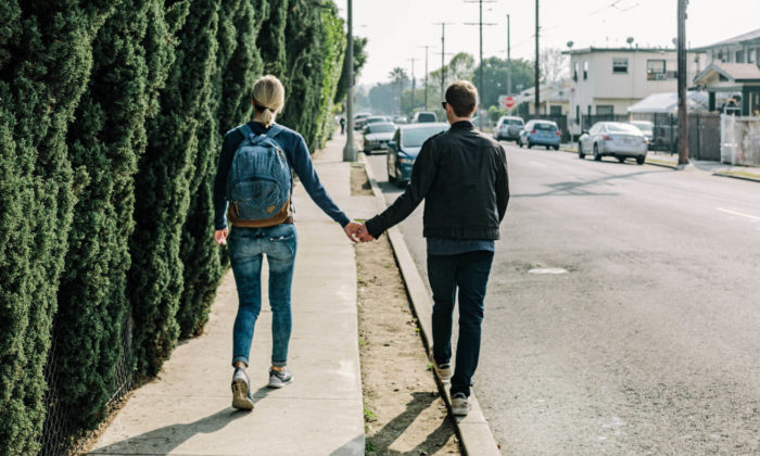 Woman and man walking down the sidewalk holding hands. Woman is wearing a denim backpack and walking in the center of the sidewalk while the man is walking on the curb of the street.