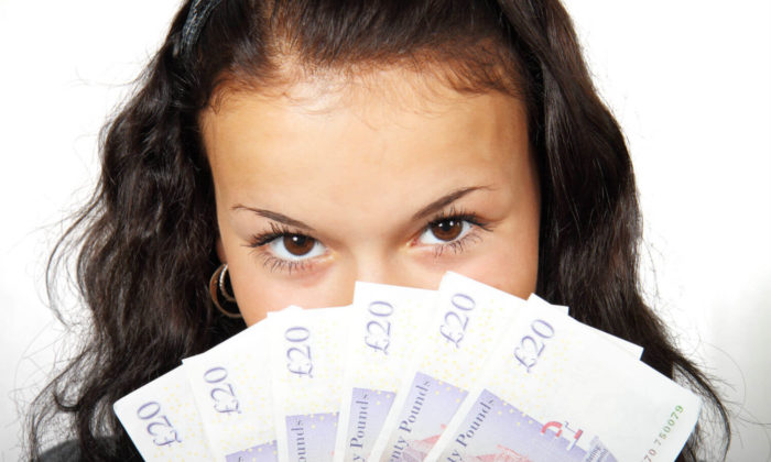 Woman hiding mouth behind a fan of money. She has long brown hair and she is making eye contact with the viewer.