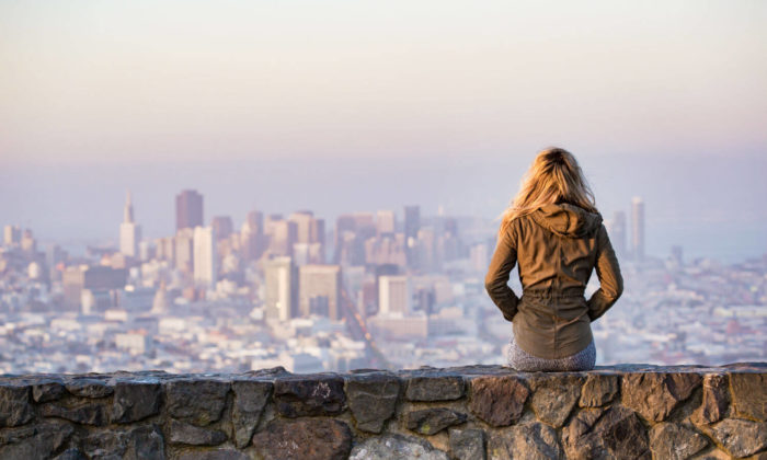 Woman in a jacket sitting on a brick wall over looking a large city scape.