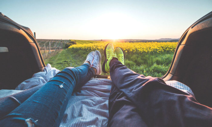 2 people sitting in the back trunk of their car, legs outstretched and a field with a sunset in front of them